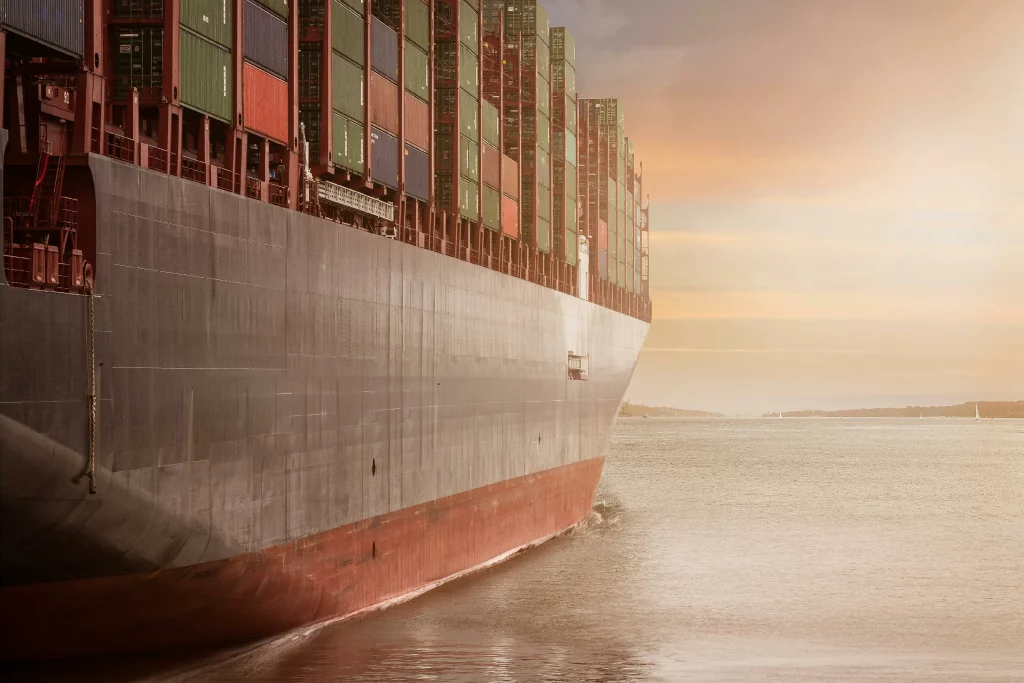 Side view of a massive cargo container ship cruising through open water at sunset, carrying tightly stacked multi-colored shipping containers.