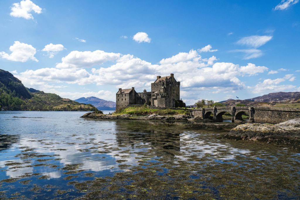 Eilean Donan Castle on a small island connected by a stone arch bridge to the mainland, with mountains reflecting in Loch Duich under a blue sky.