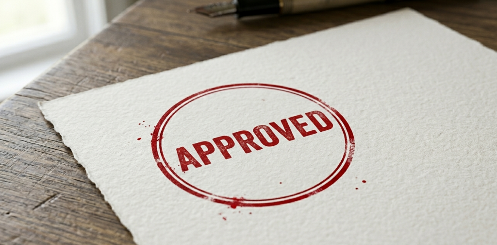 Red circular ink stamp reading APPROVED on a textured white paper document resting on a wooden desk.