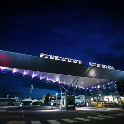 The illuminated main entrance gate and security building of Pinewood Studios at dusk under a deep blue sky.